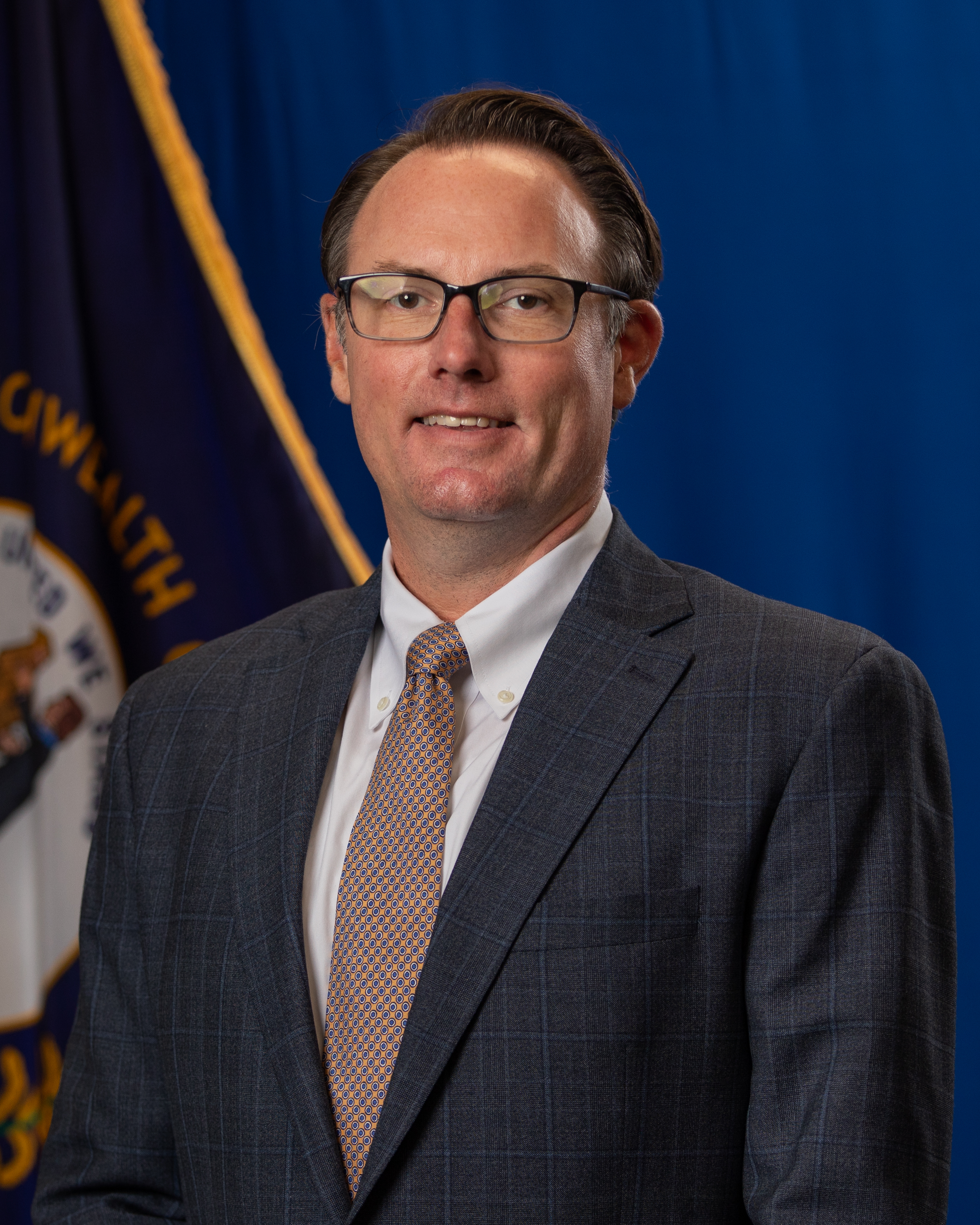 DLG Commissioner Sawyers standing in-front of a blue blackground with a State of Kentucky flag drapped on the left hand side